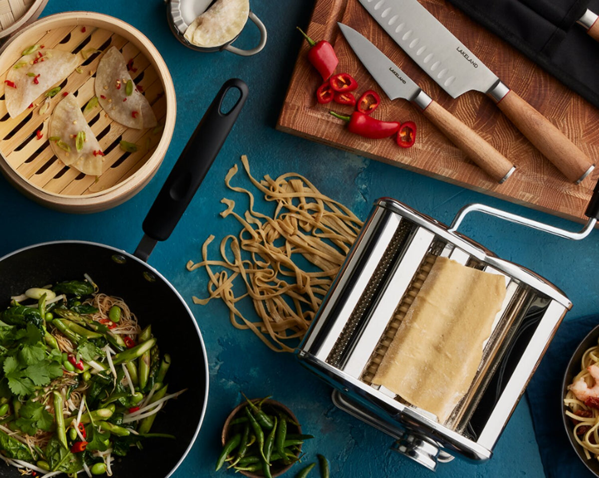A flat lay of Asian-inspired cooking tools and dishes, including a wok with stir-fried vegetables, fresh noodles, a bamboo steamer with dumplings, a pasta machine, and a set of kitchen knives on a blue background.