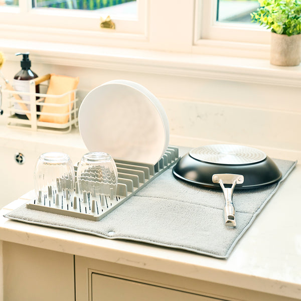 Two glasses, two plates and a frying pan are drying on a Lakeland drying mat next to the sink.